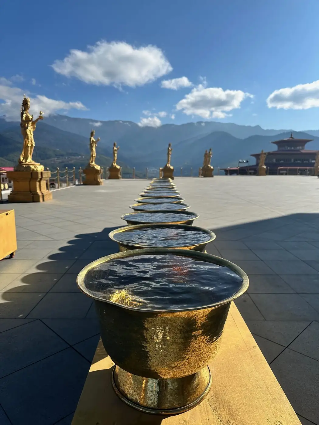 Row of golden offering bowls beneath gilded Buddha statues with mountains beyond
